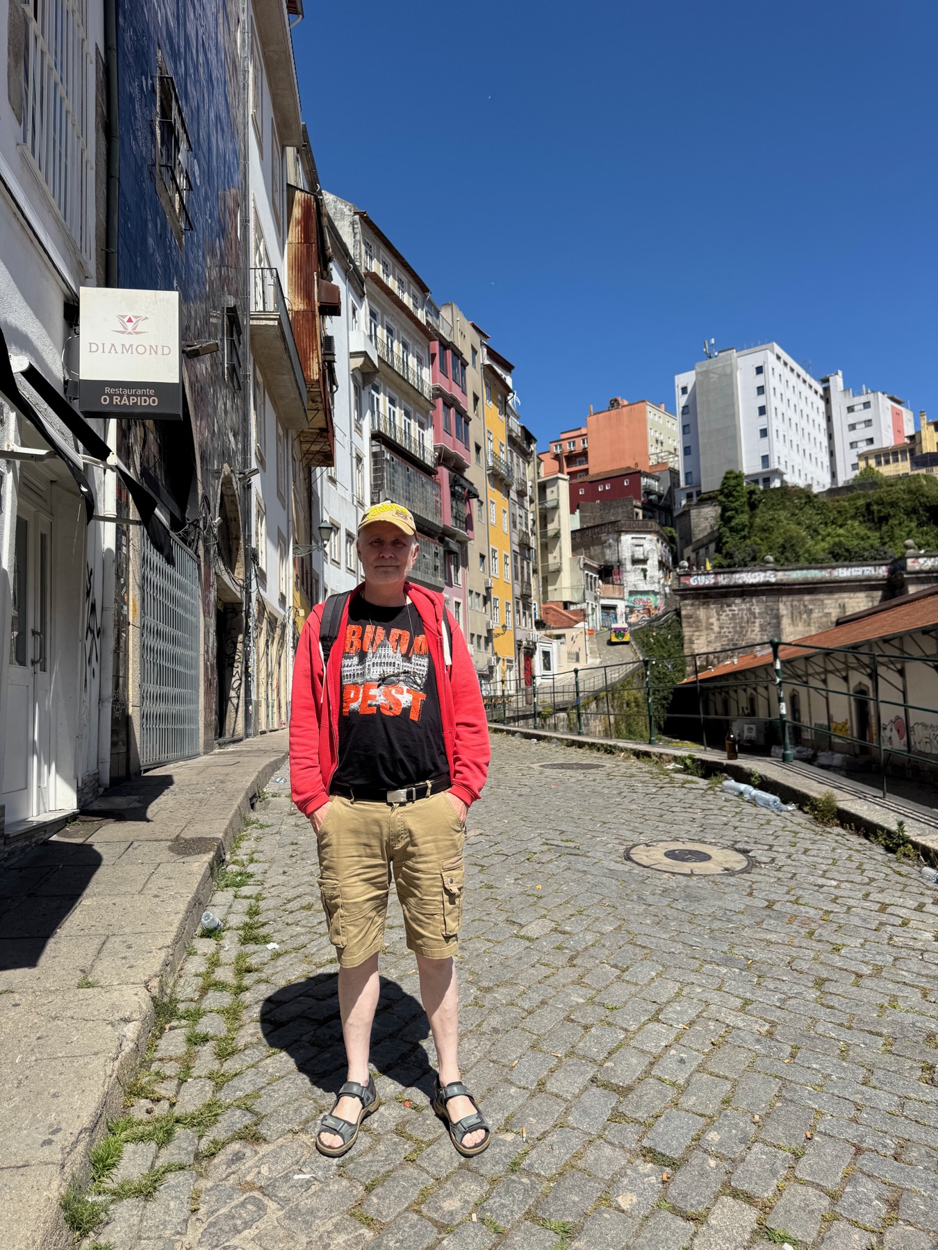 Faren i gul kasket, rød hættetrøje og Budapest-T-shirt på en solrig brostensgade.